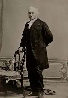 Full-figure image of a white-haired, fair-skinned man, standing with his hand on the back of a chair, wearing a dark mid-Victorian business suit