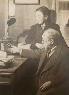 An older white man, seated at a desk, with an Asian woman standing behind him; he may be adjusting a radio dial. She is wearing glasses and a dark jacket.