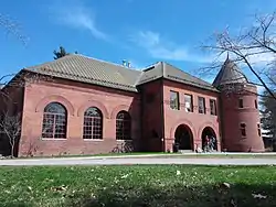 A two-story brick building with rounded windows and a tower stands on a sunny day under a mostly clear blue sky.