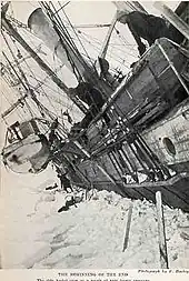  Side of a wooden steamship held in solid ice, leaning steeply to the left with a lifeboat swinging in its davits. One man visible on the ice, another aboard the ship, looking down.