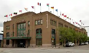 A large, rectangular, tan brick building with green trim and a roof lined with flags