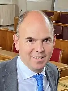 White man wearing grey suit, white shirt and blue tie