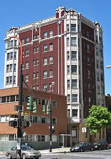 An ornate eight-story brick buildings with stone corners and trim. In the foreground is an intersection with traffic signals and some smaller buildings