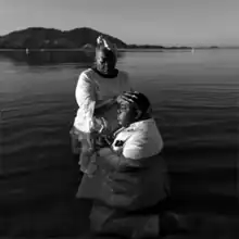 A black-and-white photograph of a woman holding a man's head in a baptism-like fashion, with both in waist-high water.