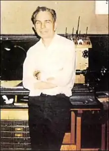 Color photograph of man leaning against a worktable with his arms crossed, a lathe, carpentry table and various tools visible behind him; he is approximately 55–60 years old with a greying head of hair and half-smile on his face.