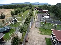 Elevated view of a series of huts and vehicles exhibited in the open air on either side of a stretch of concrete patrol road, with a range of forested hills visible in the background.