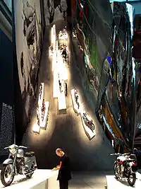 A man standing between two vintage motorcycles on pedestals surrounded by curving, reflective walls.