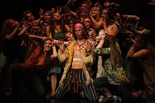 Young stage performers grouped around young white man, clean-shaven, with long hair and exposed torso