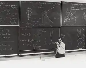 Feynman standing before a large blackboard with chalk writing all over it