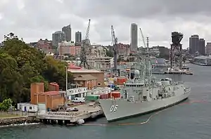 Colour photo of two grey-painted warships moored alongside wharfs. A large crane and several buildings are visible behind the ships.