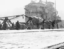 The flight deck of an aircraft carrier during a snowstorm, with the ship's superstructure in the background. A propeller aircraft is sitting on the snow-covered flight deck, and is being worked on by several people.