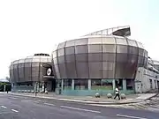 View of two of the four stainless steel drums from across an empty road. The main entrance is visible between the two drums with a circular sign above containing the words "the HUBS".