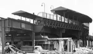 A black-and-white, slightly-angled view of a one-story brick and terra cotta Beaux-Arts building with the words "Rapid Transit" in all-capital letters over its main entrance, while the sides of the building are being demolished. Above the building are elevated railroad tracks accompanied by an island platform to the viewer's left and a curved side platform to the viewer's right. Both platforms have hipped-roof canopies, with the side platform's canopy bulging out to the right along with the platform. The side platform also has noticeable lattice railings and gooseneck light fixtures. A truck passes by on the street in front of the building.