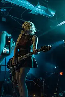 Heather Baron-Gracie holds a guitar while singing into a microphone during a concert.