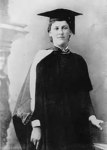 Black and white photograph of woman standing, wearing motor and gown, with book in her left hand.