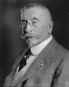 Head and torso of an older white man with a Van Dyke mustache and short hair. He is wearing pince-nez glasses and a checkered suit coat over a vest and dark tie.