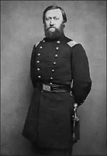 Black and white photo shows a bearded man standing. He wears a dark military uniform with two rows of buttons.