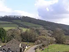 A hill covered in trees and green fields, with a house next to the road in the foreground.