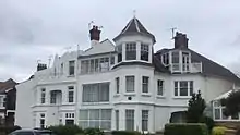 A large white house, partly with pitched roofs, and one section being surmounted by a tower-like structure with windows set in.