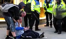 Tomlinson sitting on the ground facing the police with his arms outstretched; two men are helping him up