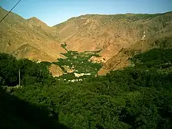 The village of Imlil in its wooded valley, surround by arid mountains. Taken early on a summer's morning, facing west. To the right a mobile phone mast sits incongruously on a nearby hill. Terracing of the valley is visible behind the village. Tall green trees are in the foreground.