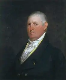 A stern-looking man with thinning, white hair wearing a white shirt and black coat with gold buttons