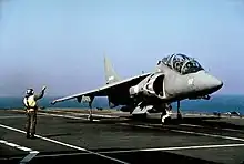 Front view of two-seat grey jet fighter on aircraft carrier deck. A directive personnel is close by.