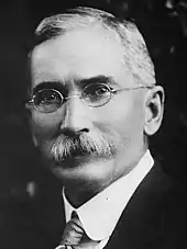 Black and white photograph of a man with a prominent mustache sporting a suit and glasses.