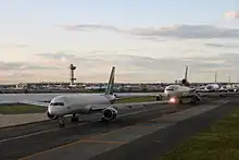 Five jumbo airplanes wait in a line on a runway next to a small body of water at John F. Kennedy Airport.