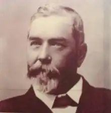 Black-and-white portrait photograph of Thallon with a moustache and goatie beard, wearing a dark suit, white shirt and dark bowtie under the collar.