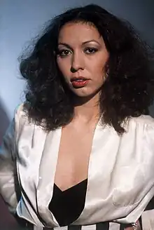 A dark-haired woman wearing a white robe with a black bustier underneath stares at the camera.