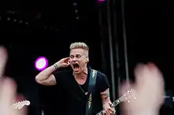 Jimi Constantine wearing a black t-shirt, standing onstage with a guitar, mouth open wide mid-vocal