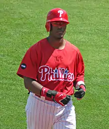 A dark-skinned man in a red baseball jersey and red left-handed batting helmet walks on a baseball field; he appears to be in his mid-twenties. His jersey reads "Phillies" in white and red script, with two blue starts dotting the "i"s.