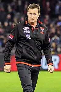 A man with light brown hair in a black jacket walks across a grassed playing field