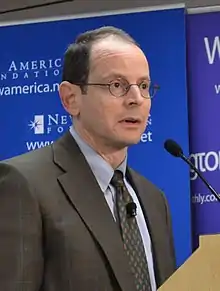 Rauch wearing a brownish grey plaid suit and round-rimmed eyeglasses, standing at a podium