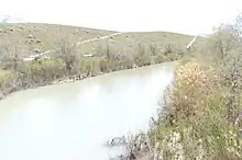 A river with plants on either side. A hill rises on the right side.