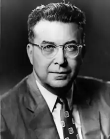 Middle aged, clean-shaven man with tousled dark hair, round spectacles and a suit