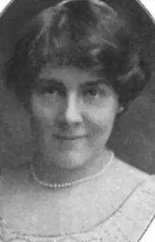 A smiling white woman with dark hair, wearing a strand of pearls and a dress with a deep scooped neckline