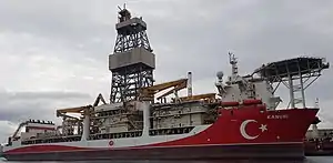 Medium-sized ship viewed from starboard. Painted mostly red with the Turkish crescent and star in white near the bow. Above the bow is a helicopter deck and there is a large derrick amidships. Two small cranes are built into the side of the ship.