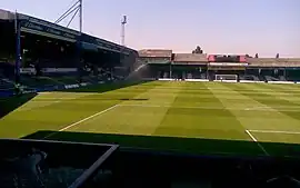 An old-fashioned association football stadium. On the left a large wooden grandstand is visible, filled with blue seats; straight ahead, a smaller stand is seen, also with blue seats. On the latter stand, the word "LUTON" is spelled out in white seats among the blue.