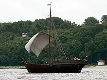 A photograph of a small wooden ship with white sail traversing an estuary; behind the ship is a wooded shoreline.