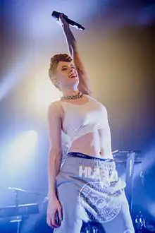 Kiesza wearing a white crop top and grey sweatpants, standing onstage with her left arm raised, holding a microphone