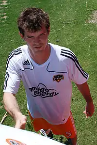 A young man with dark hair stands on a grassy surface.  He is wearing a white T-shirt with black trim and orange shorts, and is smiling slightly.