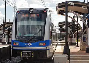 A blue and gray train stopped at a side platform station with station black and gray station canopies visible.