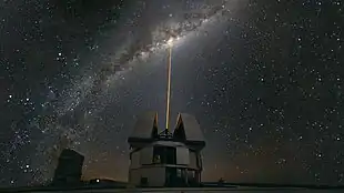 In the foreground, an astronomical observatory emits a ray of light vertically. In the background is a cluster of stars in the night.