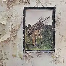 On a rough wall hangs a coloured photo of an elderly man in a field with a large bundle of sticks tied to his back.