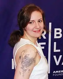 Lena Dunham in a white tank top posing in front of a step and repeat.