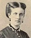 Photographic portrait of a young lady with light-colored hair swept back and wearing a high-necked, Victorian era striped dress, dark earrings and a dark locket suspended around her neck on a ribbon