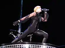 A blond woman dressed in black sings to a microphone. Behind her, a saddle can be seen.