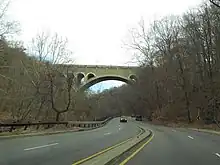 A four-lane road with a low concrete median snakes through a wooded area and under a large stone arch bridge.